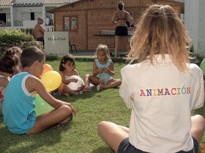 Betreuerin mit T-Shirt 'Animación' sitzt mit Kindern im Kreis auf einer Wiese am Hotel.