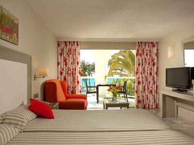 Helles Hotelzimmer mit Doppelbett, rotem Sessel und Blick auf Balkon mit Palmen.