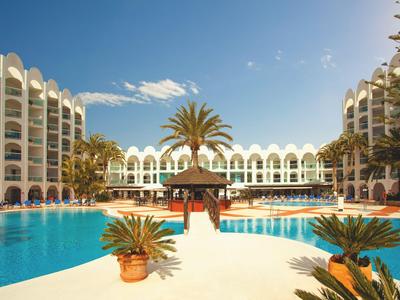 Großes Hotel mit Pool, Palmen und Sonnenterrasse unter blauem Himmel.