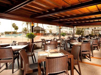 Offener Speisebereich mit Holztischen und -stühlen, Blick auf einen Strand und Palmen.
