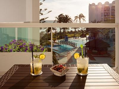 Zwei Cocktails und eine Schale Nüsse auf einem Holztisch mit Blick auf einen Pool und Palmen bei Sonnenuntergang.