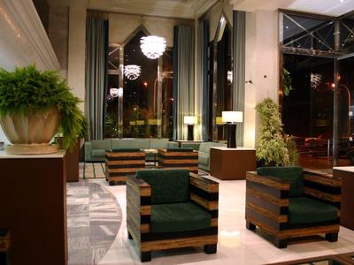 Modern eingerichtete Hotellobby mit grünen Sesseln, großen Pflanzen und hellen Lampen.