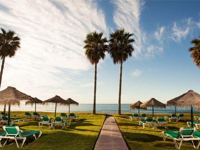 Grüner Rasen mit Liegestühlen und Sonnenschirmen vor Blick aufs Meer bei blauem Himmel.