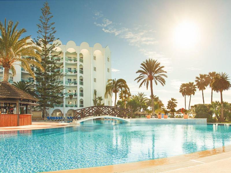 Großer Pool mit Brücke und Palmen vor einem Hotelgebäude bei Sonnenuntergang.