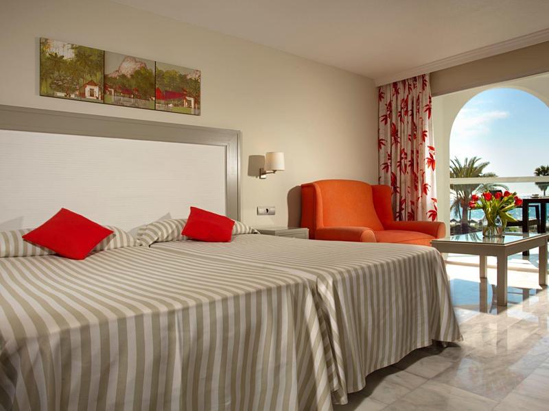 Helles Hotelzimmer mit zwei Betten, rotem Sessel und Balkon mit Meerblick.