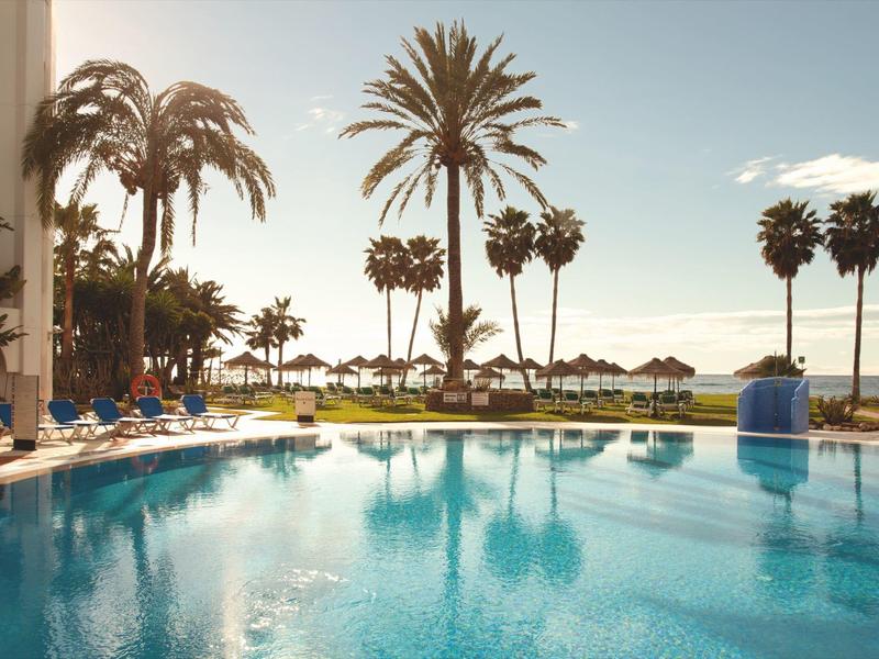 Schwimmbad mit Liegestühlen, Palmen und Meerblick bei Sonnenuntergang