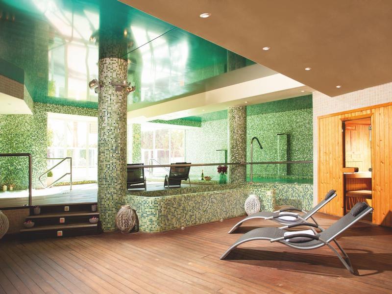 Modernes Indoor-Schwimmbad mit grünen Fliesen, Holzverkleidung und Liegestühlen im Wellnessbereich.