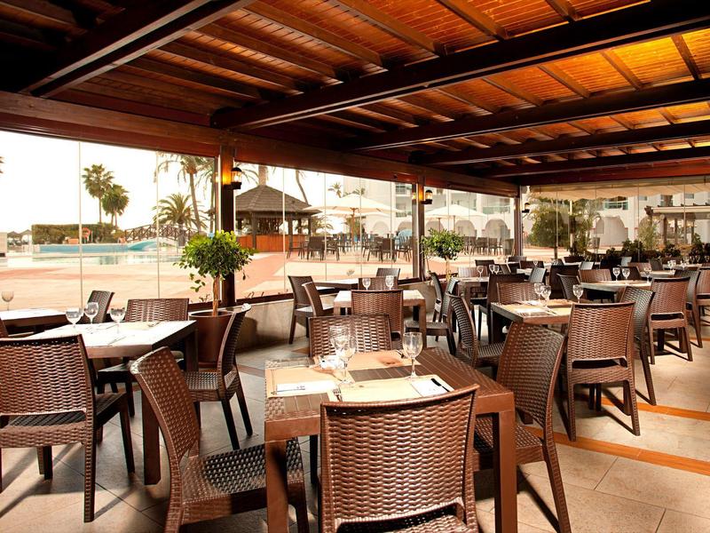 Offener Speisebereich mit Holztischen und -stühlen, Blick auf einen Strand und Palmen.