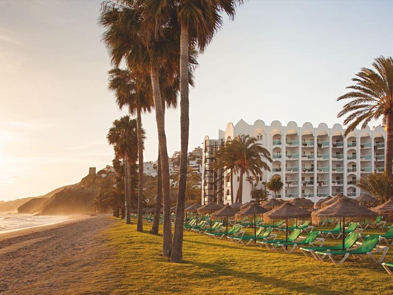 Strand mit Palmen und Liegestühlen vor einem Hotel bei Sonnenuntergang.