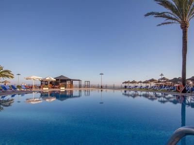 Großer Pool mit klarem Wasser, Palmen und Liegestühlen unter blauem Himmel am Strand.