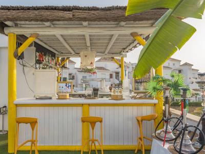 Gemütliche Bar mit Strohdach, gelben Hockern und Fahrrad vor weiß-hellgelbem Tresen.