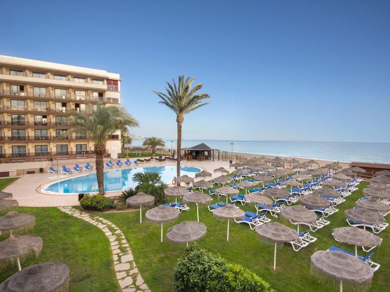 Hotel mit Schwimmbad, Liegestühlen und Sonnenschirmen auf Wiese neben Meer unter blauem Himmel.