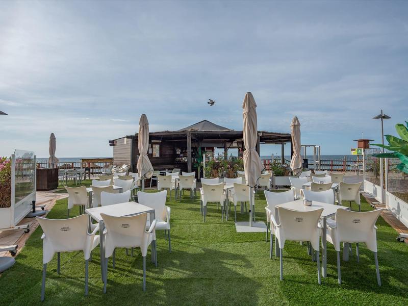Offene Terrasse mit weißen Stühlen und Tischen auf grünem Rasen, Blick auf den Himmel und Gebäude.