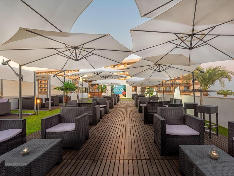 Außenbereich mit Holzterrasse, schwarzen Sesseln, Tischen und großen weißen Sonnenschirmen.