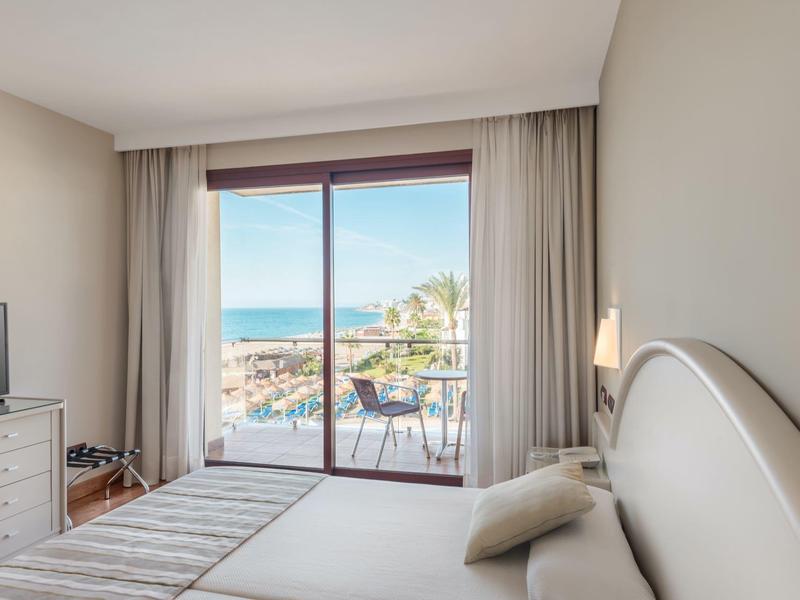 Schlafzimmer mit großem Bett, Fernseher, hellen Wänden und Balkon mit Meeresblick bei klarem Himmel.