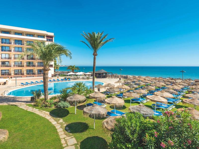 Hotel mit großem Pool, Palmen und vielen Sonnenschirmen am Strand unter klarem blauem Himmel.