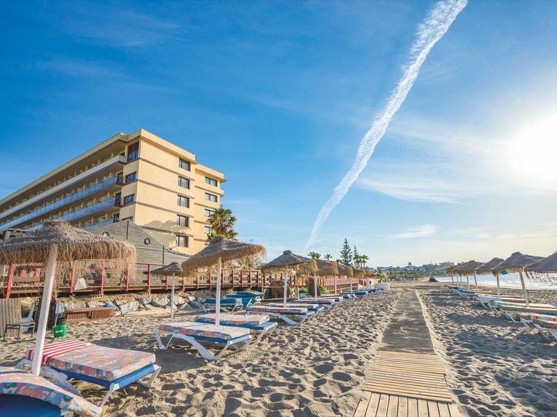 Sonniger Strand mit Sand, Sonnenliegen, Schirmen und mehrstöckigem Hotel im Hintergrund.