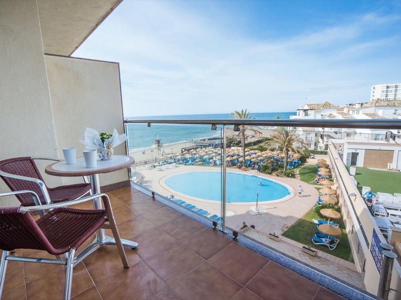 Balkon mit Tisch, zwei Stühlen und Blick auf Pool, Strand und Meer bei klarem Himmel.
