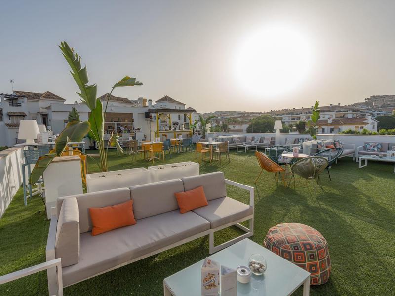 Dachterrasse mit grünen Kunstgras, Sofa mit orangen Kissen, Tischen und Pflanzen bei Sonnenschein.