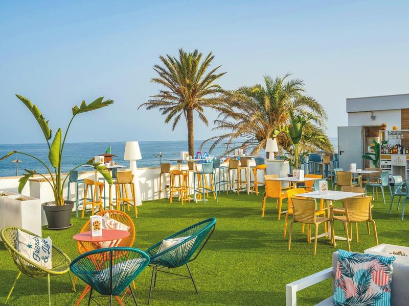 Dachterrasse mit Rasen, bunten Stühlen, Tischen und Palmen mit Meerblick im Hintergrund.