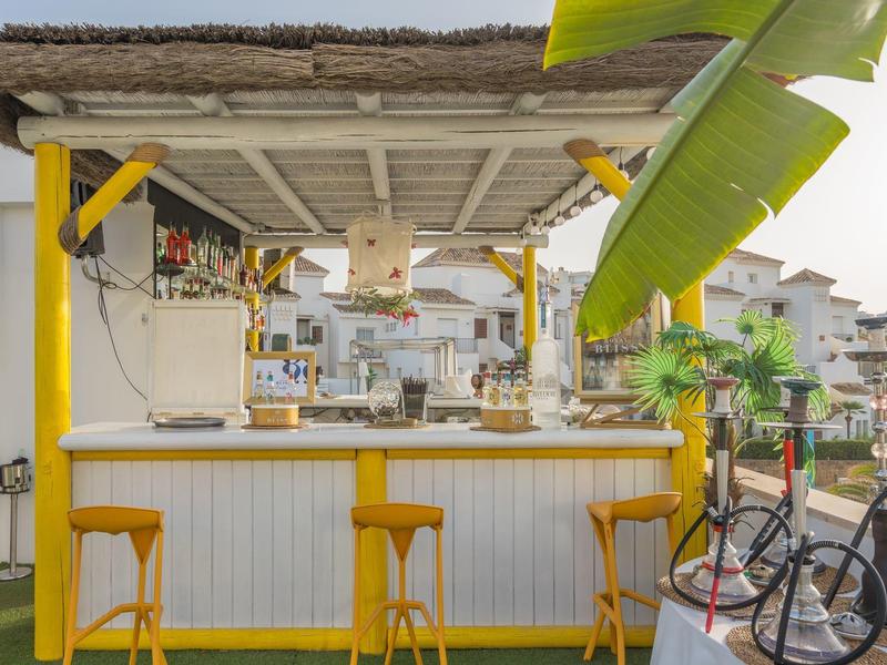 Gemütliche Bar mit Strohdach, gelben Hockern und Fahrrad vor weiß-hellgelbem Tresen.