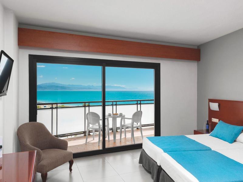 Modernes Hotelzimmer mit Doppelbett, blauen Bettwäsche, Sessel und Balkon mit Meerblick.