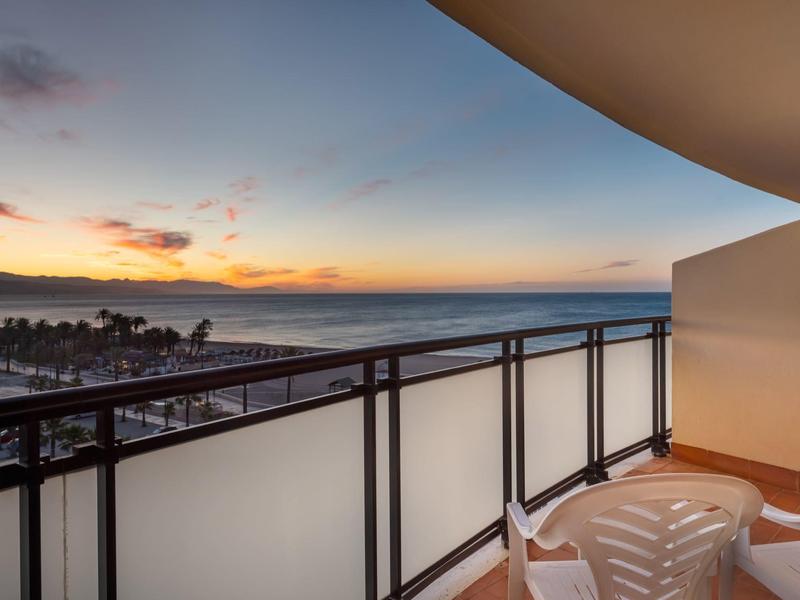 Balkon mit weißen Stühlen, Glasgeländer und Meerblick bei Sonnenuntergang.