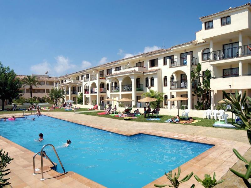 Großer blauer Pool vor mehrstöckigem, beige-weißem Appartementgebäude unter blauem Himmel.