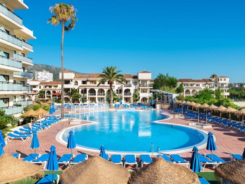 Großer, runder Pool mit blauen Liegen, umgeben von Palmen und weißen Gebäuden unter klarem Himmel.