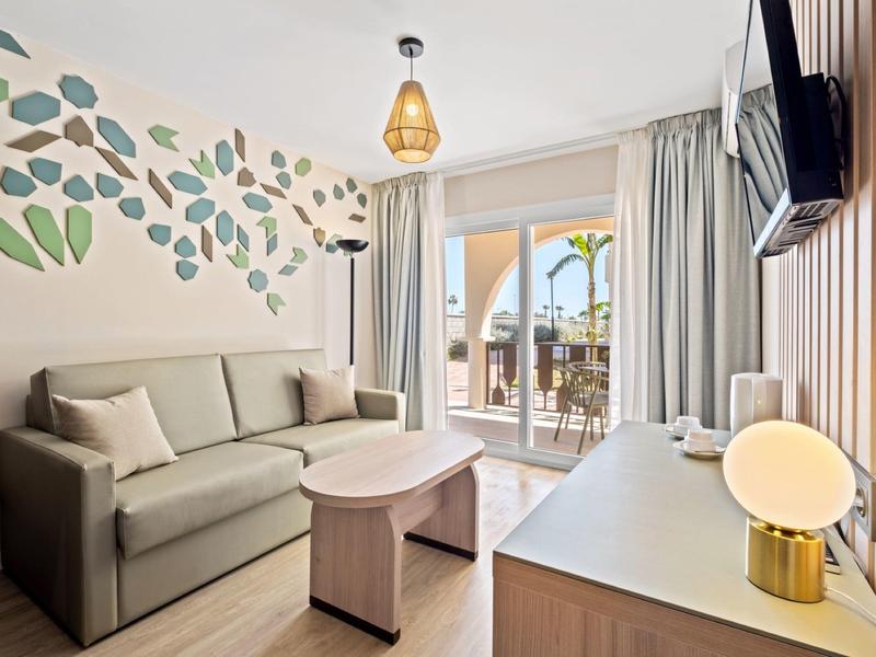 Modernes Wohnzimmer mit grauem Sofa, Holztisch, Wanddeko aus Blättern und Balkon im Hintergrund.