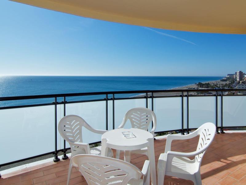 Balkon mit weißem Kunststofftisch und vier Stühlen, Blick auf das Meer bei klarem Himmel.