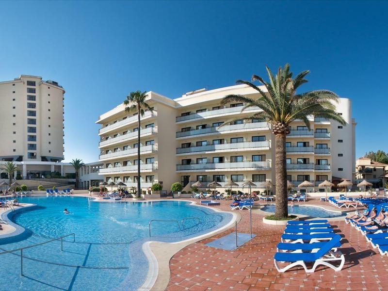 Großer blauer Pool mit blauen Liegestühlen, Palmen und mehrstöckigen weißen Hotels unter klarem Himmel.