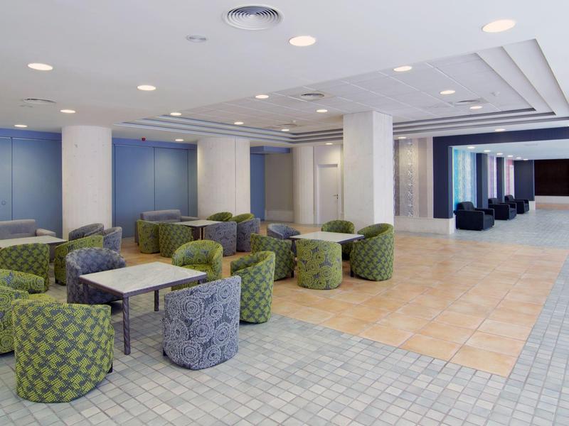 Moderner Konferenzraum mit grünen und grauen Sesseln, Tischen und großem Präsentationsbereich.