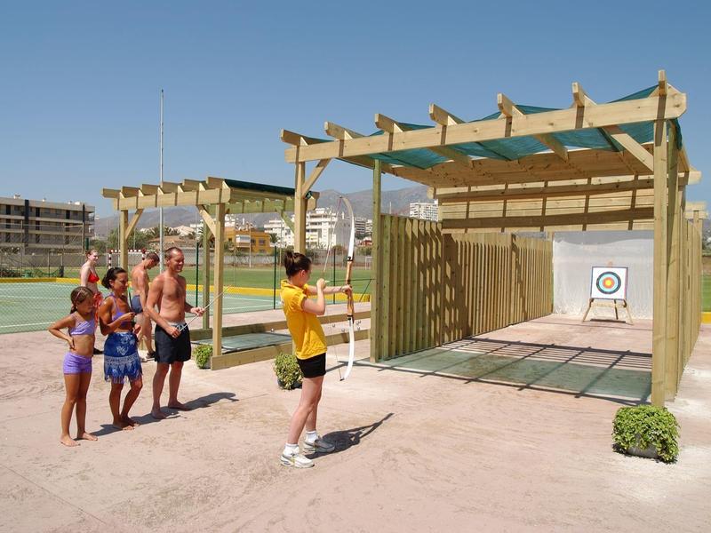 Kinder spielen auf einem Sportplatz Zielwerfen mit Wurfgeräten an einer Holzhütte.