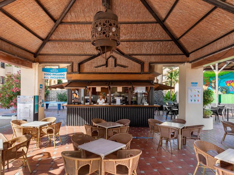 Freiluftbar mit Holzdach, Rattanmöbeln und schwarzem Tresen, umgeben von Pflanzen und bunten Wänden.