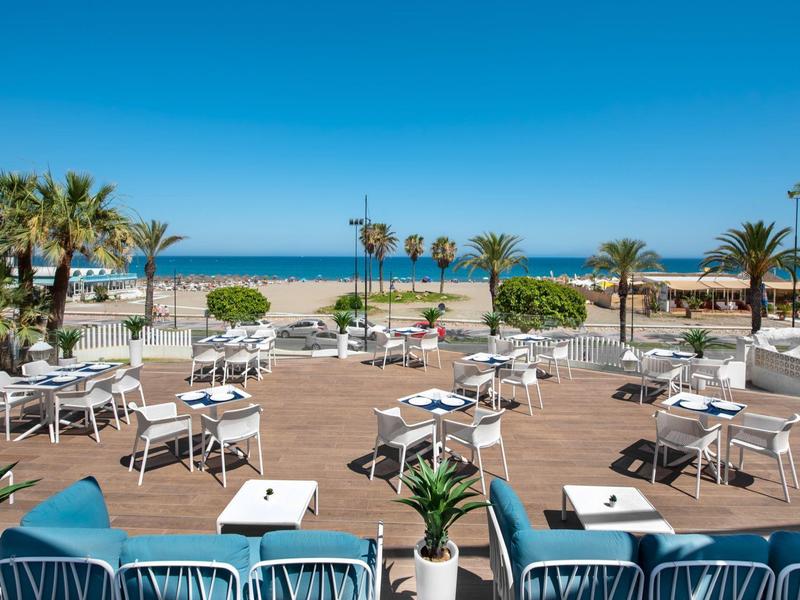 Strandrestaurant mit weißen Tischen, Stühlen, Palmen und Blick aufs blaue Meer unter klarem Himmel.