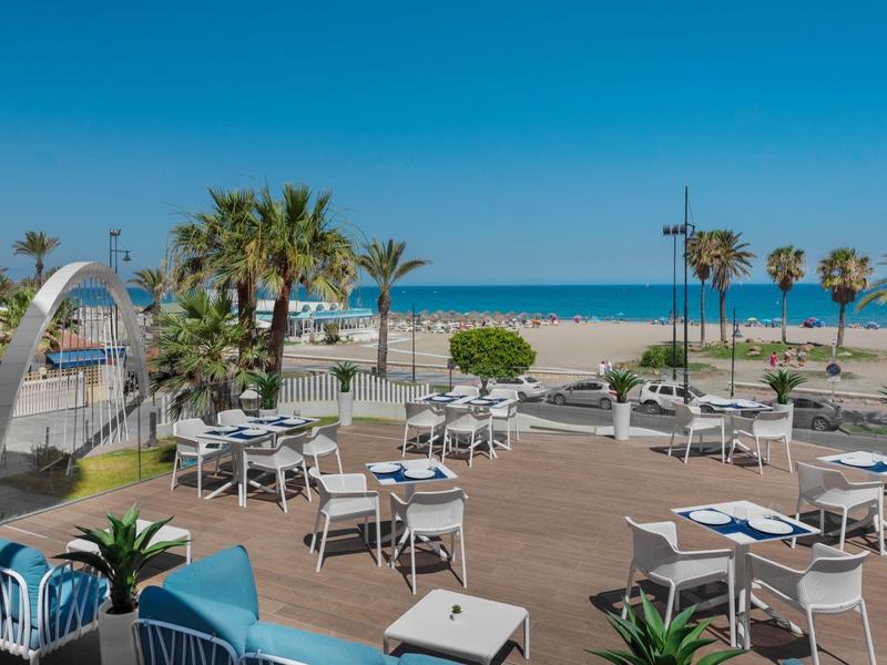 Terrasse mit weißen Stühlen, Tischen, Palmen, Sandstrand und blauem Himmel am Meer.