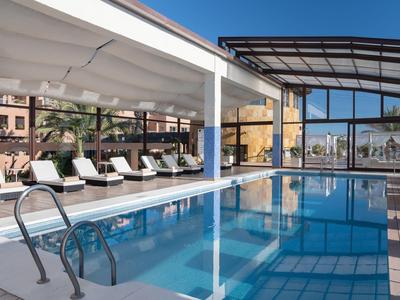 Moderner Außenpool mit Liegestühlen, Glasdach und angrenzendem Gebäude bei klarem Himmel.