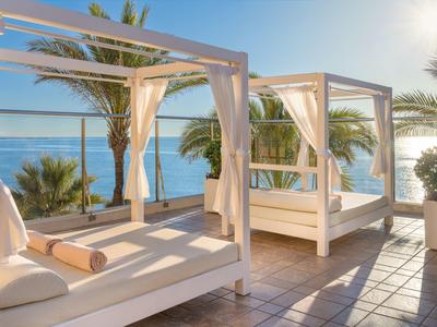 Terrasse mit zwei weißen Daybeds, Palmen und Blick auf das blaue Meer bei Sonnenschein.