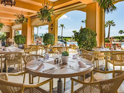 Offenes Restaurant mit runden Tischen, Korbstühlen und Blick auf Palmen und blauen Himmel.