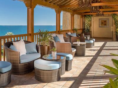Überdachte Terrasse mit Korbmöbeln, blauen Kissen, Pflanzen und Meerblick bei klarem Himmel.