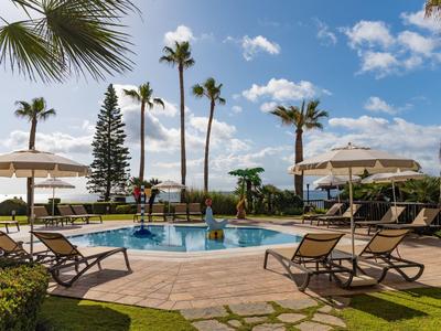 Schwimmbad mit Liegestühlen, Sonnenschirmen und Palmen unter blauem Himmel mit weißen Wolken.
