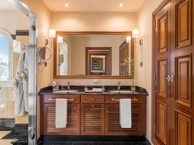 Hotelová koupelna s dvojitým umyvadlem, velkým zrcadlem, dřevěnými skříňkami a jasným osvětlením.