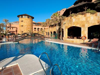 Großer Pool mit blauer Wasserfläche umgeben von mediterranen Gebäuden und klarem Himmel