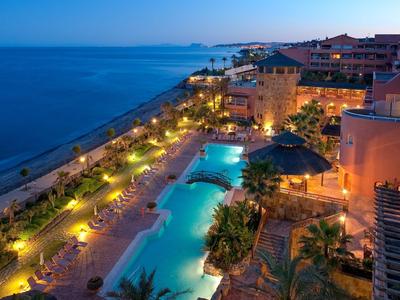 Blick auf einen beleuchteten Hotelpool und Strand bei Nacht mit ruhigem Meer im Hintergrund.