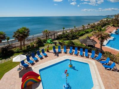 Hotelpool mit Kinderbereich und Meerblick, umgeben von Liegestühlen und Palmen.