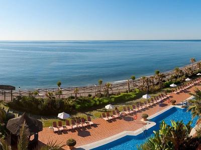 Panoramablick auf Küste mit Pool, Palmen und Sonnenliegen bei klarem Himmel.