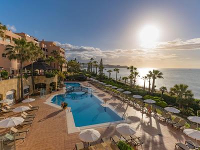 Sonniger Poolbereich mit Liegestühlen, Palmen und Meerblick bei hellem Himmel am späten Nachmittag.