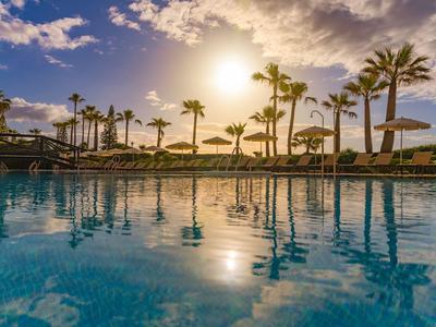 Schwimmbecken bei Sonnenuntergang mit Palmen und Sonnenschirmen am Ufer.