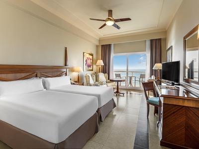 Modernes Hotelzimmer mit zwei Doppelbetten, Schreibtisch, Fernseher und Balkon mit Meerblick.